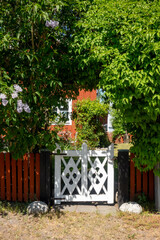 White wooden gate on falu red paint fence leading to a garden below lush green trees on each side...