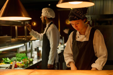 Young serious female worker of restaurant in uniform preparing food in the kitchen while standing...