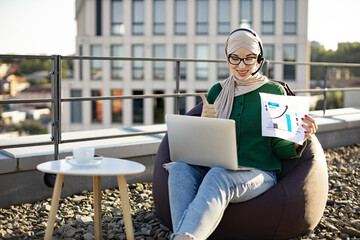Focused muslim adult pointing at paper infographics during online meeting held outdoors via modern technologies. Calm businesswoman in hijab discussing financial data plan on panoramic terrace.