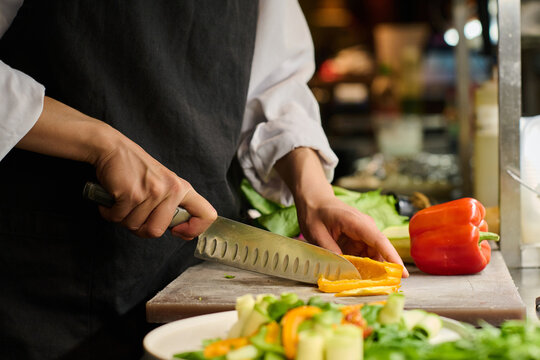 Close-up of hands of young female kitchen worker chopping fresh yellow pepper for salad or some other dish while standing by workplace