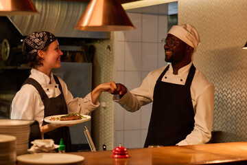 Two happy young intercultural kitchen workers in uniform giving each other punch bump while one of them holding plate with beef steak