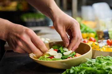 Hands of chef or kitchen worker putting small slices of fresh mozarella cheese on chopped green lettuce before mixing ingredients of salad