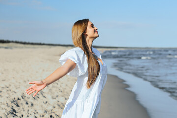 Happy smiling beautiful woman is on the ocean beach in a white summer dress, open arms