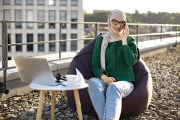 Fototapeta premium Relaxed muslim lady in headscarf smiling at camera while talking on cell phone near small table with laptop outdoors. Charming employee exchanging information with client about project outside office.