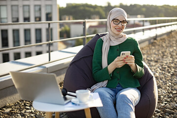 Close up of cheerful person with smartphone resting in pouf chair on roof terrace during coffee break on midday. Happy arabian female in hijab and eyewear texting messages via mobile in open air.