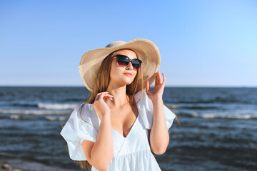 Happy blonde woman is posing on the ocean beach with sunglasses and a hat. Evening sun