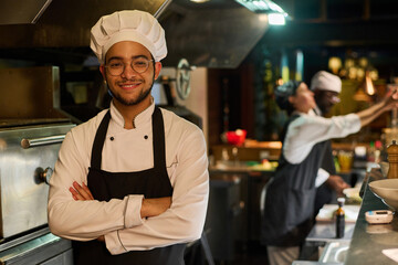 Young smiling cook in uniform keeping his arms crossed by chest and looking at camera while...