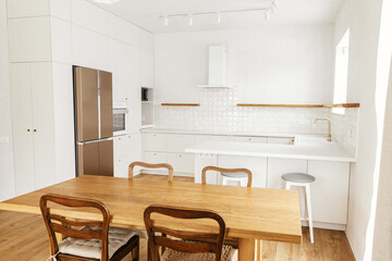 Modern kitchen interior. Stylish white kitchen cabinets with brass knobs, granite counter, appliances and rustic wooden table in new scandinavian house. Modern minimal kitchen design.