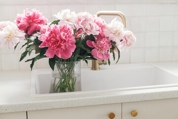 Beautiful peonies in vase in sink on background of brass faucet and white counter in new scandinavian house. Pink peony flowers in modern kitchen interior, summer floral arrangement