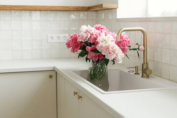 Beautiful peonies in vase in sink on background of brass faucet and white counter in new scandinavian house. Pink peony flowers in modern kitchen interior, summer floral arrangement