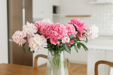 Beautiful peonies in vase on wooden table on background of stylish white kitchen with appliances in new scandinavian house. Modern kitchen interior and summer floral arrangement