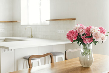 Beautiful peonies in vase on wooden table on background of stylish white kitchen with island, wooden shelves and appliances in new house. Modern kitchen interior in farmhouse