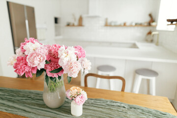 Modern kitchen interior. Beautiful peonies in vase on wooden table on background of stylish white kitchen with appliances in new scandinavian house.Summer floral arrangement