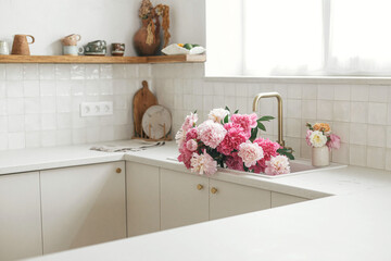 Pink peony and roses flowers in modern kitchen interior, summer floral arrangement. Beautiful peonies in sink on background of brass faucet and white counter in new scandinavian house