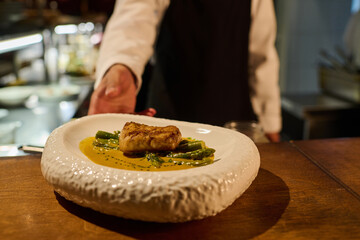 Male chef in uniform taking plate with tasty roasted fish fillet with green beans and sauce while going to serve it to guests of restaurant