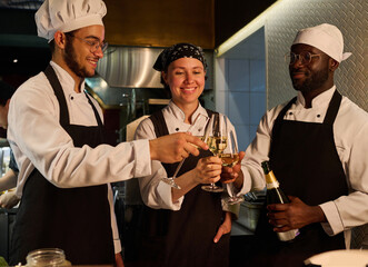 Three happy young intercultural cooks celebrating life event with champagne in the kitchen while toasting and clinking with flutes