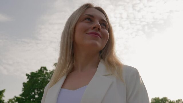 Portrait of a Beautiful Blonde Woman Smiling at the Camera. Attractive Woman with a Natural Beauty of the Face in a White Jacket on the Background of a City Park. Slow Motion.