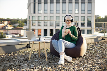 Young smiling female wearing hijab and headphones holding mobile device while resting in soft chair on rooftop. Happy arabian adult checking list of tracks before enjoying music during lunch break.