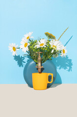 Beautiful natural daisy flowers and yarrow in a vase with golden faucet and yellow cup. Minimal concept of refreshment. Frontal view.