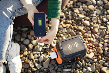 Top view of female holding smartphone with full charge on screen being plugged into portable power station via cable. Young woman keeping digital device running using off-grid electricity in open air.
