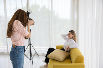 An Asian experienced professional woman photographer in her captivating studio shooting the beautiful woman model sitting in a sofa. Discover the fusion of fashion, style.