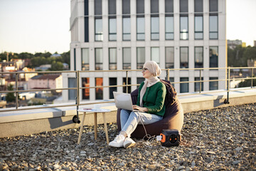 Wide view of arabian woman in traditional headscarf working on laptop while sitting in soft chair...