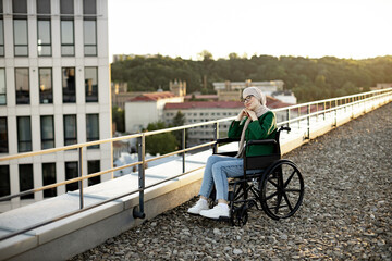 Pretty bespectacled woman with mobility issues daydreaming with hands under chin in sunlight on roof terrace. Relaxed muslim person wearing jeans and hijab soaking up with vitamin D outdoors.