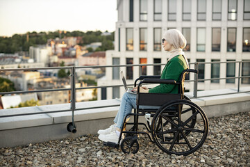 Obraz premium Side view of muslim lady in headscarf using digital device for remote work on panoramic terrace. Well -organized businesswoman in wheelchair searching information over internet application outdoors.