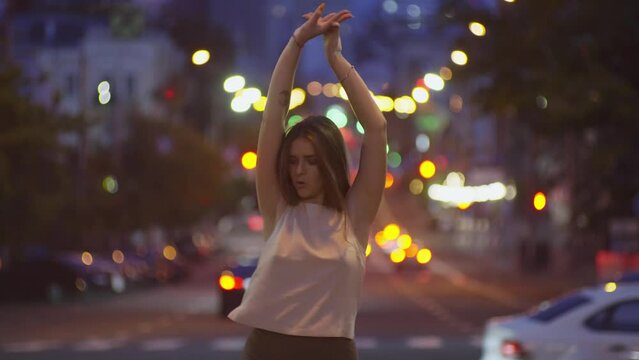 A beautiful Caucasian girl is dancing against the background of city lights. Urban landscape woman stomps music and moves to the rhythm of the dance. Lifestyle video. High quality 4k footage