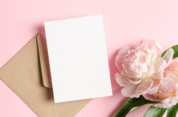 Fototapeta premium Blank invitation or greeting card mockup with envelope and pink peony flowers, top view, copy space
