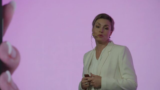 A woman presenter is standing on a well-lit stage in front of a large LED screen showing a hand holding a generic smartphone with green screen. Technology, application or product launch presentation 