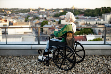 Charming muslim lady in traditional clothing with cell phone in hands while unwinding on roof terrace. Calm bespectacled woman in wheelchair relaxing while taking in bird's-eye view of city.