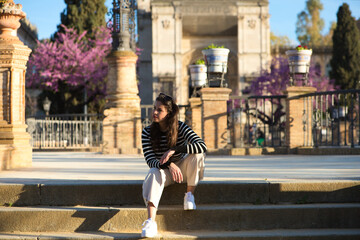 Beautiful young woman with long brown hair wearing a black and white striped T-shirt and white trousers, sitting on the steps of a square in a park in Seville.