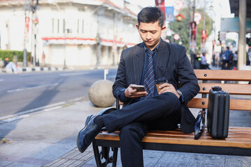 Fototapeta na wymiar Professional millennial Asian businessman in a formal business suit looking at mobile phone sitting outside on city street with copy space