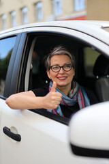 An adult woman in glasses is driving a white car and showing a thumbs up. Successful car rental or...