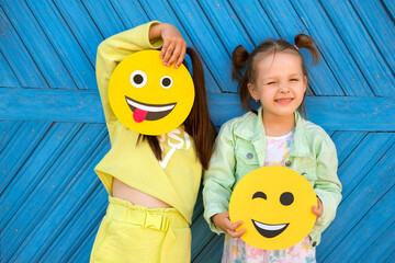 Humorous funny girls hold various funny smiley faces and make faces standing at the blue wall - obrazy, fototapety, plakaty