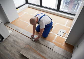 Top view of man in workwear using metal construction ruler and pen while drawing line on laminate...