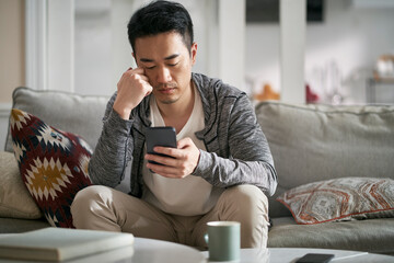 young asian adult man sitting on family couch at home looking at cellphone
