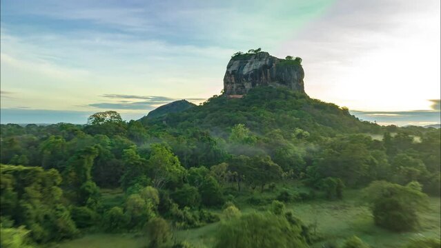Forwards fly above wet paddy fields and wild nature around rock with steep walls. Hyperlapse footage of ancient rock fortress against colourful morning sky. Sigiriya, Sri Lanka