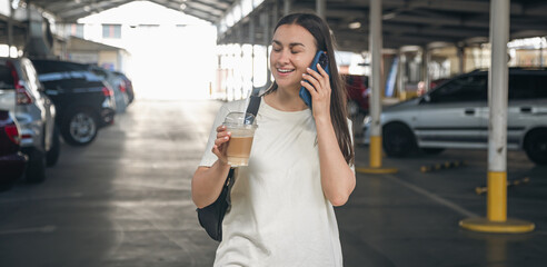 Fototapeta premium A young woman with coffee talking on the phone in the parking lot.