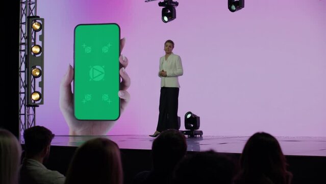 A woman presenter is standing on a well-lit stage in front of a large LED screen showing a hand holding a generic smartphone with green screen. Technology, application or product launch presentation 