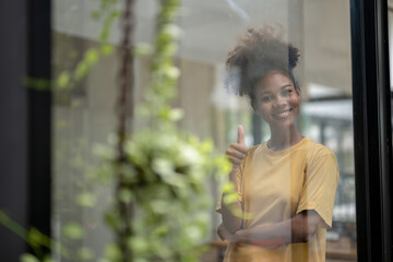 Shot from outside of young African American woman standing thoughtful in modern office looking out of window. thumb up to photographer.