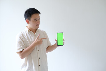 An Asian man wearing a beige shirt is pointing and looking to a smartphone with green screen that held by left hand. Isolated white background. Suitable for advertisement.