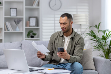 Young hispanic businessman working remotely at home on laptop. Holding the phone and anxiously looking at documents, checking accounts, financial problems and bankruptcy