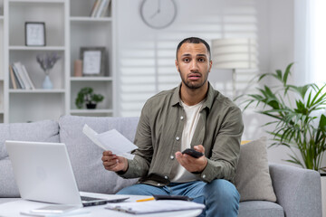 Portrait of a worried young African-American man sitting on the sofa at home, holding a phone and documents. Checks accounts, has financial problems and debt. He looks sadly at the camera