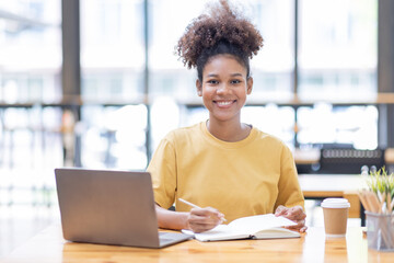 Business And Education Concept. Smiling african american  sitting at desk working on laptop writing letter in paper documents, free copy space. Happy millennial female studying using laptop	