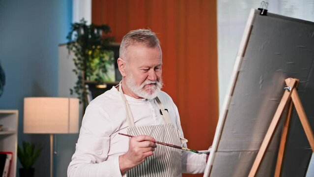 creative pensioner, elderly man is happy to paint a picture using brushes with paints and an easel at home