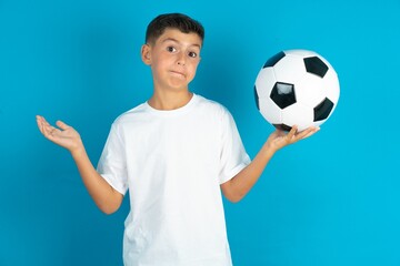 Clueless Little hispanic boy wearing white T-shirt holding a football ball shrugs shoulders with...