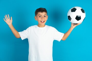 Optimistic Little hispanic boy wearing white T-shirt holding a football ball raises palms from joy, happy to receive awesome present from someone, shouts loudly,