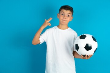 Unhappy Little hispanic boy wearing white T-shirt holding a football ball makes suicide gesture and imitates gun with hand, curves lips, keeps two fingers on temple